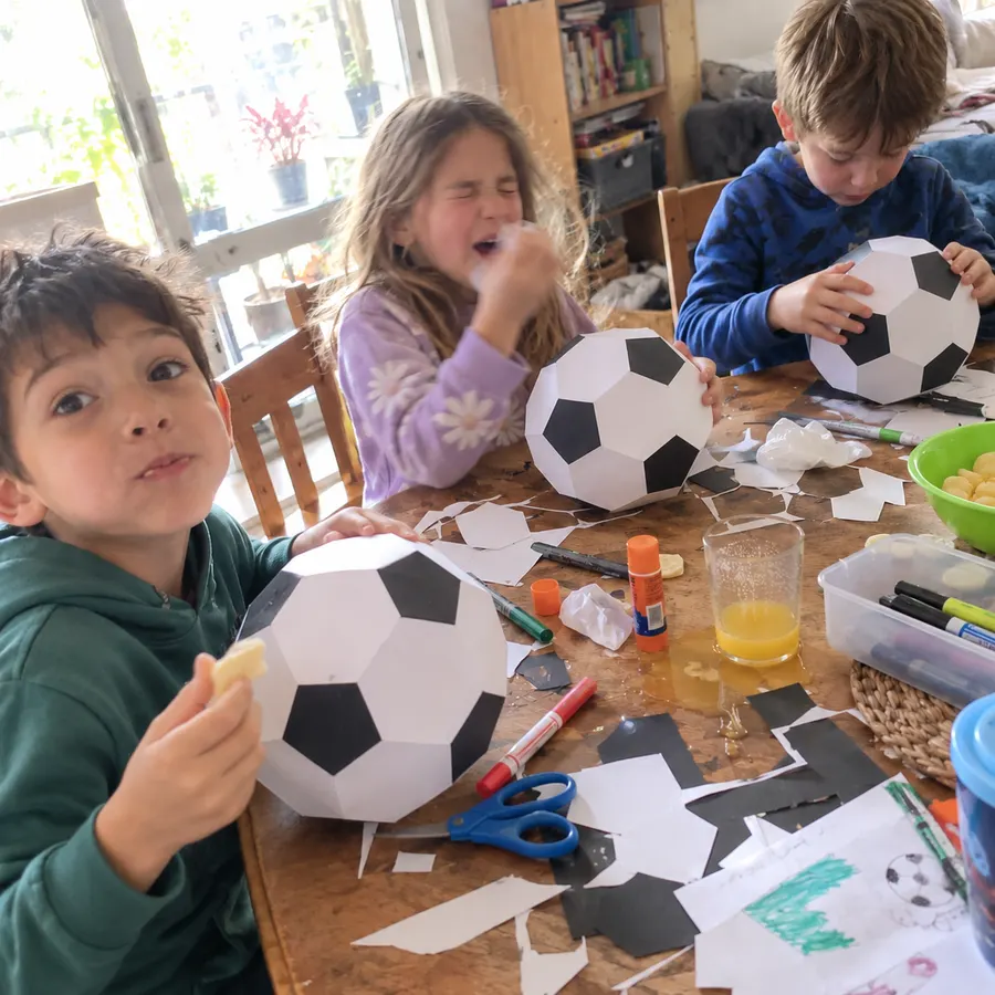 Three kids at a wooden table cutting and gluing paper footballs