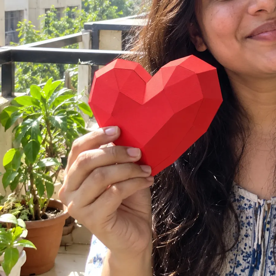 A smiling woman holding a red paper heart on a balcony