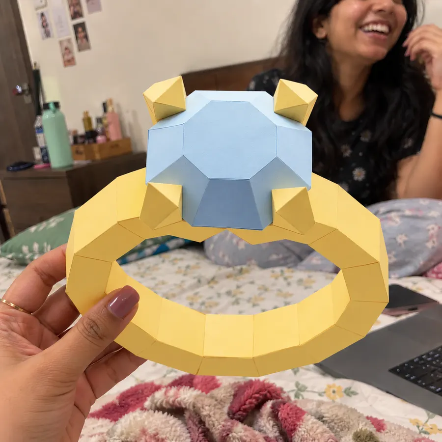 A hand holding a large yellow paper ring with a pale blue gem, a friend laughing behind